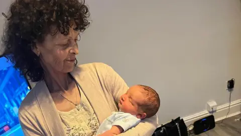 An older woman with curly dark hair smiles down as a newborn baby she is holding. 