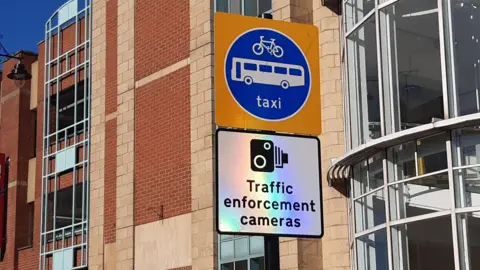 LDRS A white square bus gate enforcement sign, reading Traffic enforcement cameras. There is a second sign above it with a picture of a bus, bike and the world taxi written on it.