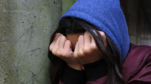 A hooded person holds their hands to their face.