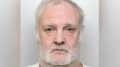 A mugshot of a man with white hair and a short white beard on his face and neck.