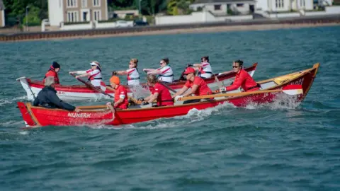 Two coastal rowing teams go head to head one in a red boat and red tops, the other in a white boat with white tops and a red stripe