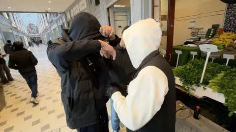 Shaun Whitmore/BBC Suspect in handcuffs covers his face while surrounded by two plain-clothed police officers in a shopping precinct.