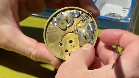 The back of the unfinised pocket watch, a gold piece with various silver components, being held by two hands. 