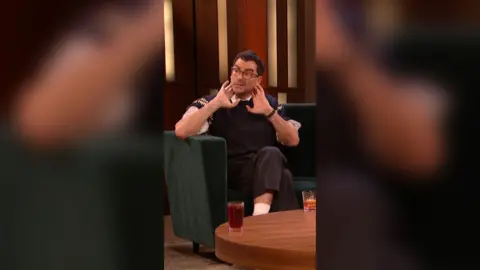 Dan Levy has dark hair, is wearing glasses and is holding his hands to his neck.