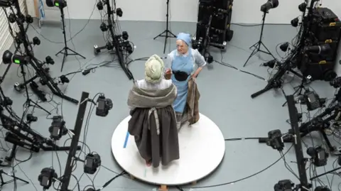 Rachel Carter Two women in period dress clasp hands while surrounded by camera equipment. The image is taken from above the women