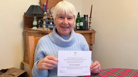 Ms Ferguson is in a baby blue knitted turtleneck jumper and is sitting at a table that has a red and green striped tablecloth. She has short white hair and is smiling. Behind her is a wooden cabinet with a blue lamp and glass bottles on it.