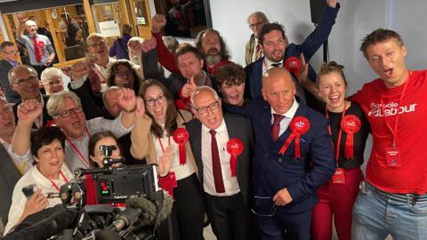 A group of about 20 people wearing red Labour t-shirts and rosettes cheering in front of a television camera