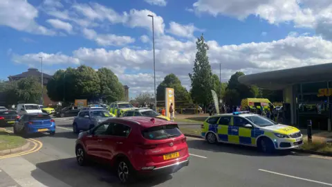 Cars driving along a main road past Waterworld in Stoke-on-Trent. A large number of police cars and ambulances are parked at the building across the street.