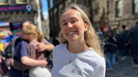 BBC Courtney Buchner. Courtney is a woman in her 20s with long blonde hair worn loose. She has a nose ring and smiles at the camera. She's pictured outside in Edinburgh on a busy street, wearing a white T-shirt. 