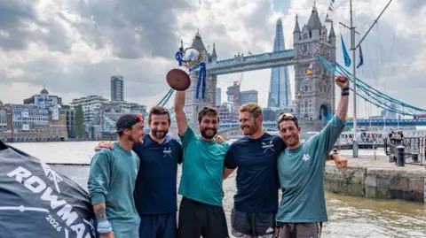 GB Row Challenge Five men standing on the banks of the River Thames, with the ancient Tower Bridge behind them, and further in the background, the towering glass Shard building. They are all wearing blue shorts and T-shirts.