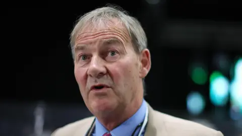 PA Media Rupert Lowe has short brown greying hair. He wears a light fawn jacket and light blur shirt with red tie.
