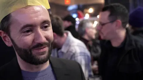 Man in blue jumper and black blazer wearing a yellow paper Christmas hat stands outside a pub