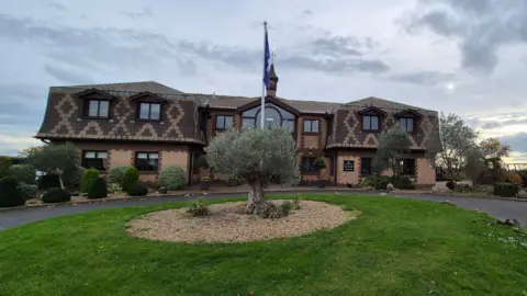 Stuart Woodward/BBC The West Essex Golf Club clubhouse building, photographed in November 2025. It is a light brown brick building with dark brown panelling over the second storey, with a diamond pattern of lighter panels within it. There are several large windows above the main entrance. There is also a large circular area of grass in front of the building with a gravel circle in the centre, as well as bushes and a flagpole with a dark blue flag hanging from the top.