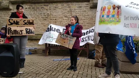 NEU A young girl stood holding a cardboard sign and a microphone. Next to her are three other people who are holding banners. All of the signs say "save our schools".