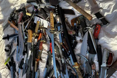 West Midlands PCC Dozens of large knives, blades and swords laid out on a white blanket.