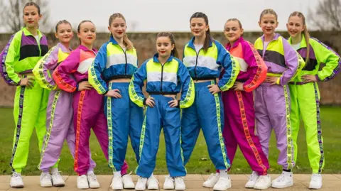 Richard Jarmy Photography A line-up of nine girls, each with hair scraped back in a ponytail, and wearing red lipstick and shellsuit-type outfits in various combinations of bright pinks, blues, purple and neon green.