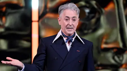 Alan Cumming on stage in a black suit talking at the Bafta Film Awards in London, 2026