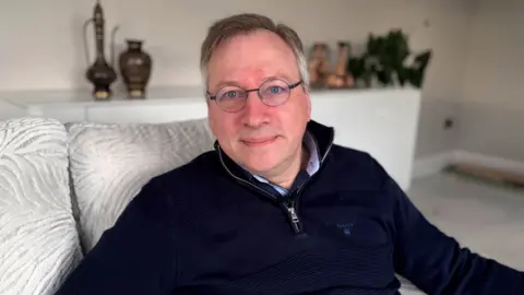 A man with round glasses and short grey hair sits on a silver sofa, looking down the lens of the camera. He has his lips pursed and his eyes are a piercing blue colour. He is wearing a navy quarter zip jumper with a silver zip.