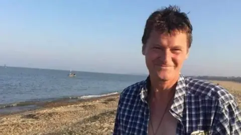 Norfolk Police Nigel Mazs standing on a beach on a hot day smiling with his shirt open.

