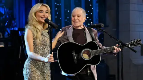 Todd Owyoung/NBC via Getty Images Sabrina Carpenter, dengan rambut pirang melengkung, senyum memegang mic dan bahu Paul Simon saat dia melihat ke arahnya mengenakan blazer beludru dan memegang gitar akustik hitam