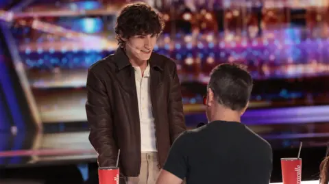 Chris Haston/NBC Magician Joel Mawhinney appears on the America's Got Talent TV show.  He has  short, curly brown hair and is wearing a brown leather jacket, beige trousers and a white shirt.  He is standing at a desk facing Simon Cowell who has his back to the camera.   Cowell has short, dark hair and is wearing a black t-shirt. 