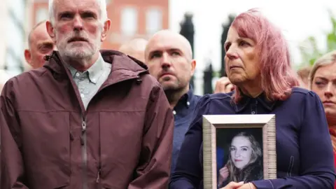 A group of solemn people stand together outdoors, with a woman in the centre holding a framed photo of a young woman while others hold hands beside her.