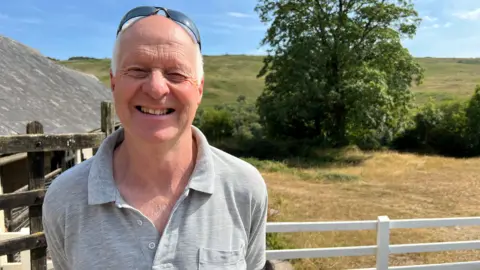 Un hombre con un polo gris y gafas de sol en la cabeza está de pie en una granja, frente a una puerta blanca, en un día soleado, sonriendo a la cámara.