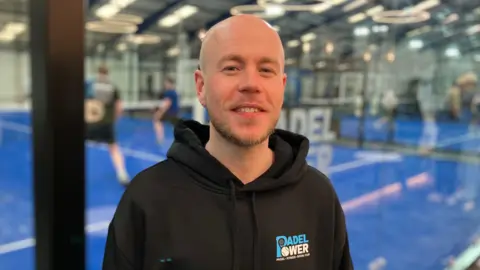 Sam Read/BBC A man in a black branded Padel Power hoodie stands against a glass panel on the back wall of a padel court where a match is being played.