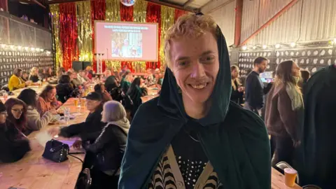 A young man with curly blonde hair smiles at the camere while wearing a dark gren cloak. In the background are long tables with customers sipping drinks.