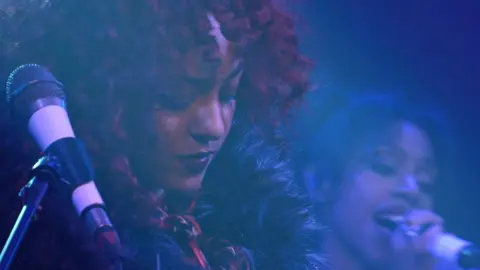 John Fairhall/BBC Nova Twins perform on stage. A close-up photo of Georgia South, who has dark red curly hair, she is looking down at a guitar out of shot, and has a microphone next to her. Over her shoulder is Amy Love, who is singing into a microphone.