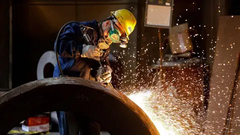 Getty Images A person wearing a bright yellow helmet, goggles and a mask covering their lower face works in a factory amid a shower of sparks