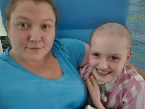 Family A woman and a young girl hug in a blue chair. The woman has brown hair, and is wearing a nose ring and a blue T-shirt. The girl has no hair and is wearing a pink and white striped top. She has a small plastic tube and device attached to the front of her neck.