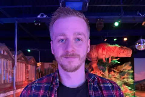 Stefan Edwards is looking straight at the camera. He has a moustache and beard and is wearing a checked shirt over a black top. He is standing the middle of the arcade with a dinosaur seen behind him