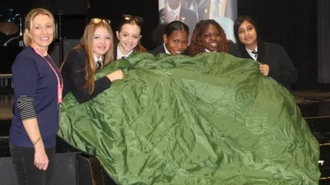 Lawn Manor Academy A lady is standing smiling to camera in a school hall next to a group of five female students who are all huddled under an open green sleeping bag