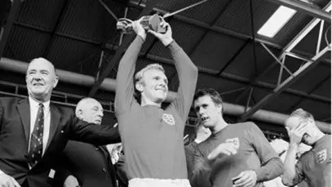 Captain Bobby Moore, who is wearing his football kit, lifts the World Cup trophy in the air in the stadium surrounded by players.