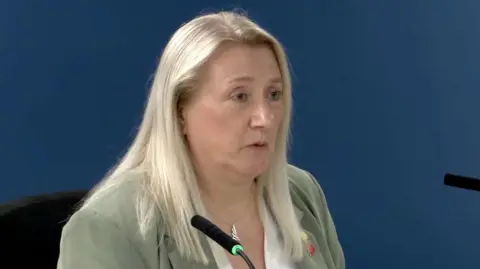 A woman with long blonde hair wearing a green blazer with a white shirt. She has a long silver necklace and is speaking into a mic at the inquiry.