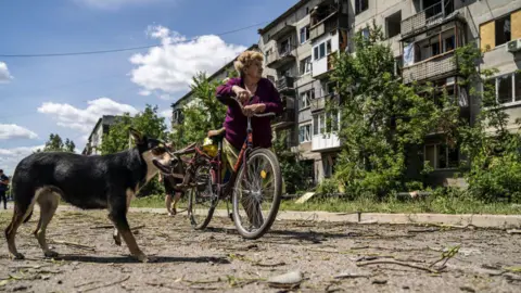 Getty Images Một người phụ nữ dựa vào xe đạp gần một con chó, nhìn lên một tòa nhà bị hư hại