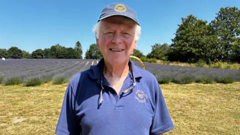 BBC A man in a navy polo shirt with a yellow round logo with heart shape stands in front of a field of lavender. He is wearing a navy cap with the same circular logo.