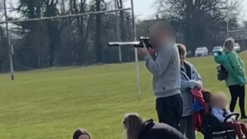 BBC A man in a grey jacket at a pitch side looking down a barrel-like fitting on the device on a football pitch.
