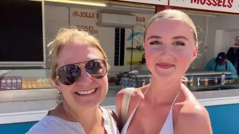 Dionne Howell and her daughter Poppy standing close as they both smile at the camera. Dionne is wearing a white linen top and sunglasses while Poppy is wearing a white bikini top. They stand in front of a stall. 