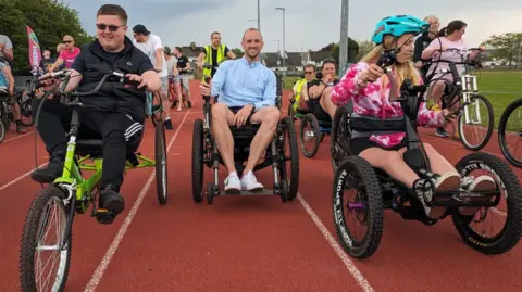 People gather on an athletics track to try out different adaptive bicycles and tricycles. People taking part are smiling.