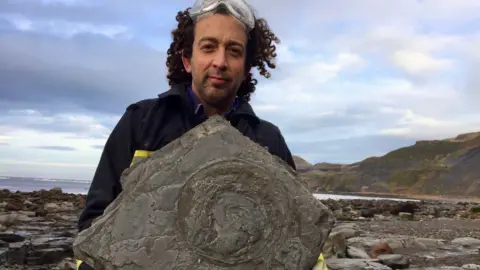 Steve Cousins/www.YorkshireCoast.rocks A man with dark curly hair holds up a large rock with the imprint of a fossil in the centre. He has clear plastic glasses on the top of his head, and is dressed in dark workman-style clothes, with bright green gloves. A rocky coastline and the sea can be seen behind him.