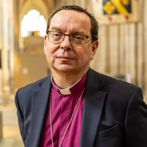 Diocese of Winchester The Right Reverend Philip Mounstephen is standing in a church, with the chain of a cross visible around his neck. He is wearing a suit and a burgundy shirt, with a white dog collar. He has glasses.
