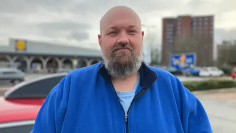 A shaven-headed man with a greying beard. He is wearing a blue fleece over a blue t-shirt, and is standing by a car in a car park