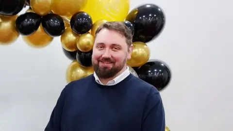 Ben Pickering Man with short brown hair and beard wears a white shirt and navy blue jumper. He is smiling and stands in front of a balloon arch in gold and black.