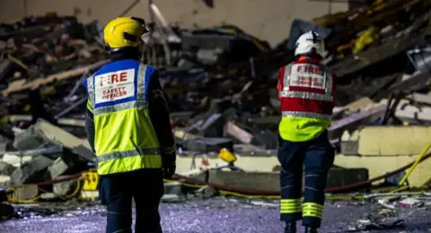 Two firefighters in hi vis look at a large pile of debris.