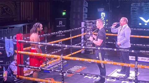 Mr Sarpong in a black boxing ring. He is wearing red shorts and gloves, and a man in all black is filming him from his phone. In the middle of the ring is an announcer, who is wearing a grey suit.