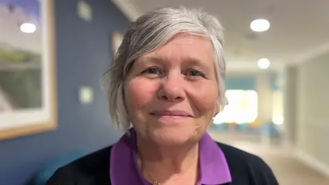 Emma Baugh/BBC Genia Haylock is standing in a corridor. She has short grey hair and is wearing a purple shirt with a black jumper over the top.