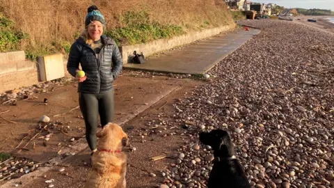 Sea swimmer Kate Gardner stands on Budleigh Salterton beach, which has large stones. Her two dogs - a golden retriever called Basil and black Labrador called Fergus - are standing in front of her. Gardner has a tennis ball in her hand. She is wearing a black padded coat over her wetsuit and a blue woolly bobble hat.