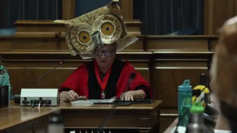 Councillor Dave Shields wearing red Lord Mayoral robes and a large owl headdress. He is sat in the council building delivering a speech to a crowd of people also dressed up in nature inspired outfits